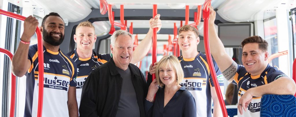 Brumbies players riding a Canberra Metro light rail vehicle with happy middle-aged couple